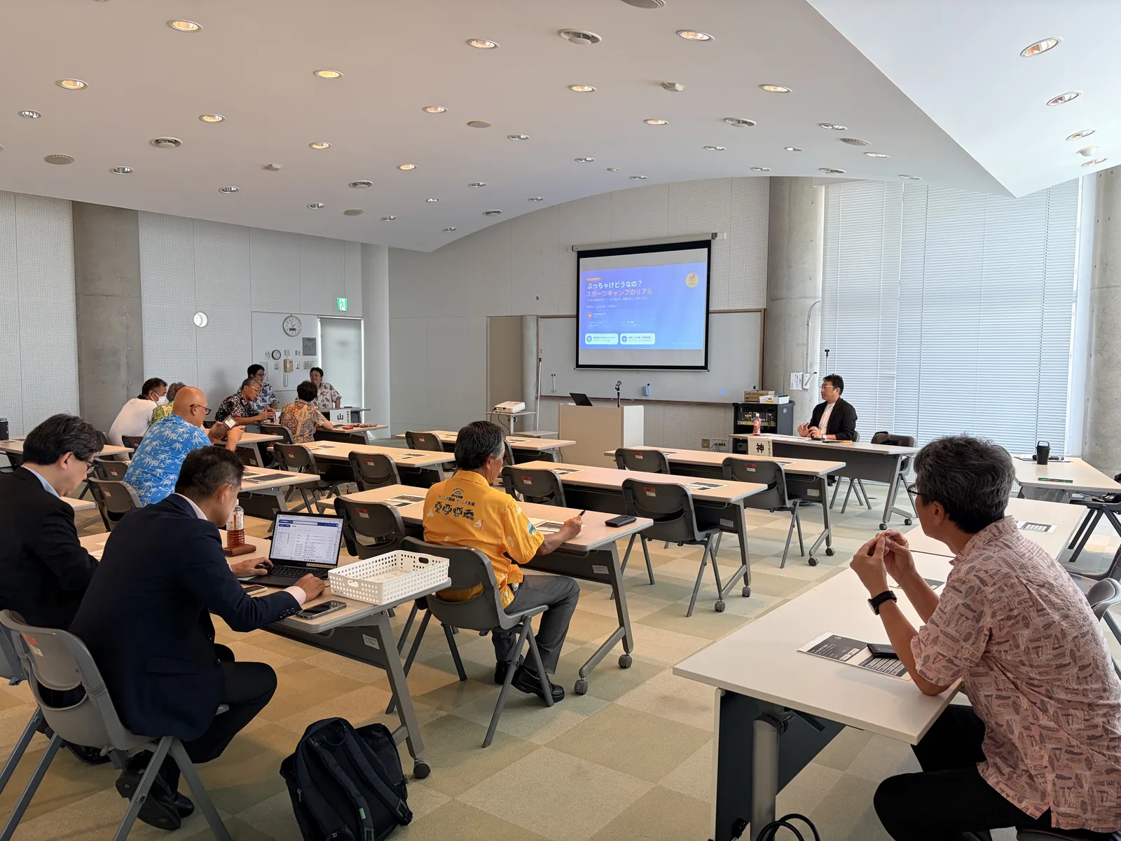 ふれあいプラザなのはな館の会議室に設営されたスクリーンとスクール形式の座席。開会前の会場の様子