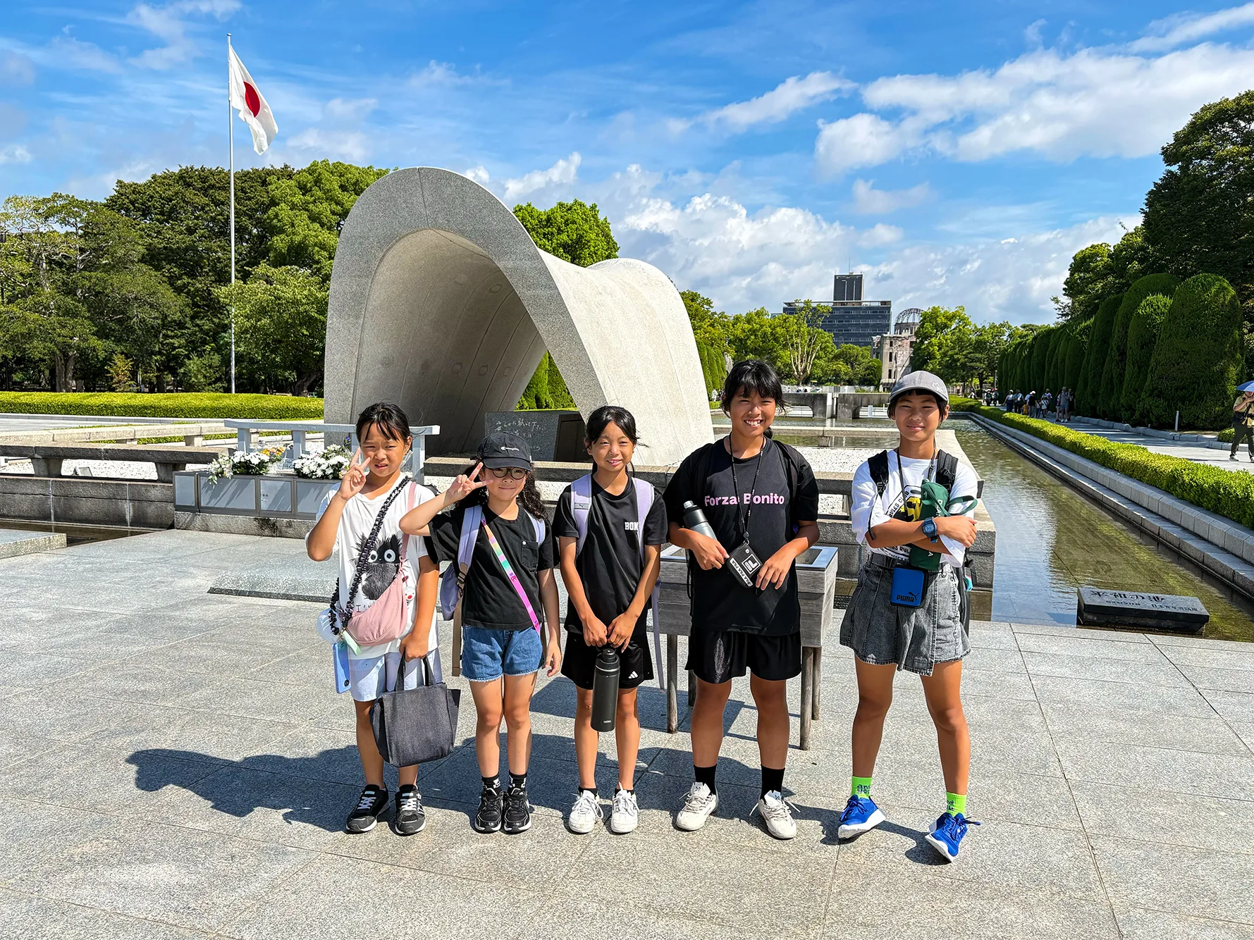 広島平和記念公園の原爆死没者慰霊碑の前に立つ子どもたち。奥には原爆ドームが見える